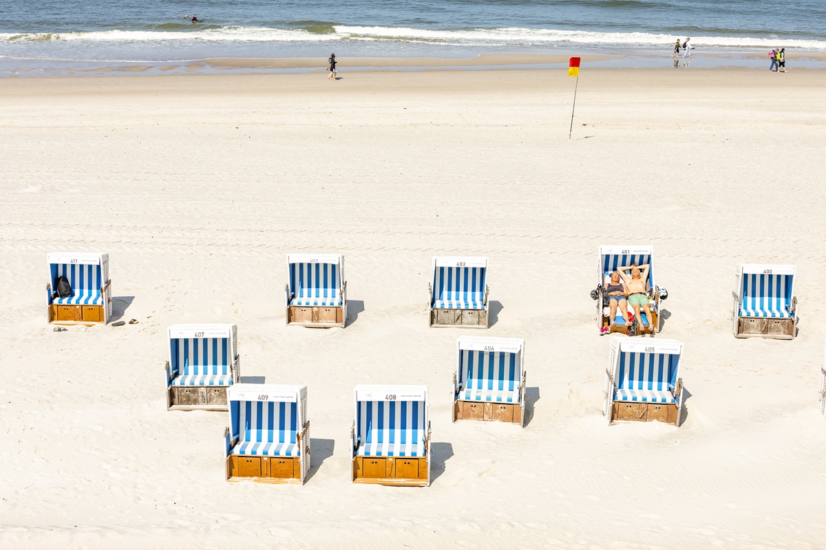 Eine Reihe von blau-weiß gestreiften Strandkörben am weißen Sandstrand von Sylt, mit Menschen, die die Sonne genießen.